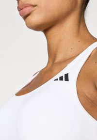 Λευκό αθλητικό σουτιέν με λαιμόκοψη scoop, λεπτές τιράντες και μαύρο λογότυπο στον αριστερό ώμο. Ομαλή, ελαστική υφή υφάσματος.