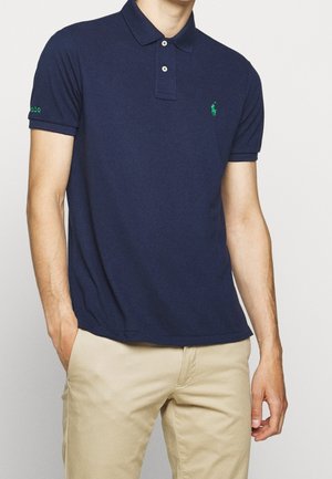 Homme portant un polo bleu marine à manches courtes avec un logo vert sur la poitrine et un pantalon beige, les mains partiellement dans les poches, sur un fond uni.