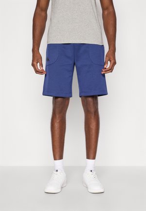 Homme portant un short bleu au niveau des genoux, un t-shirt gris, des chaussettes blanches et des baskets blanches, debout devant un fond clair uni.