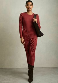 Abito lungo a maniche, di colore bordeaux, con dettaglio di ruches in vita, silhouette aderente, abbinato a una borsa a spalla marrone e stivali al ginocchio.