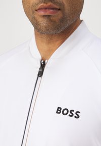 Witte zip-up jas met zwart "BOSS" logo op de linkerborst, met een gladde textuur en contrasterende ritsdetails.