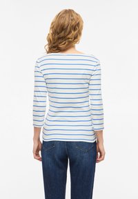 Γυναίκα με κυματιστά μαλλιά που φοράει λευκό πουκάμισο με μακριά μανίκια και μπλε οριζόντιες ρίγες και σκούρο μπλε τζιν, γυρισμένη με την πλάτη προς τα πίσω σε ένα απλό φόντο.