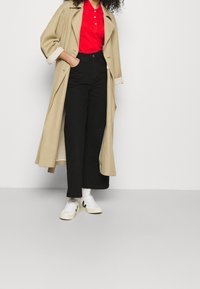 Personne portant un long manteau beige sur un polo rouge, un pantalon large taille haute noir, des chaussettes blanches et des baskets blanches avec un logo noir.