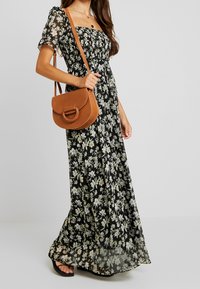 Maxiklänning i svart tyg med vit och gul blommig tryck, korta puffärmar, elastisk bodice, tillsammans med en brun axelremsväska.