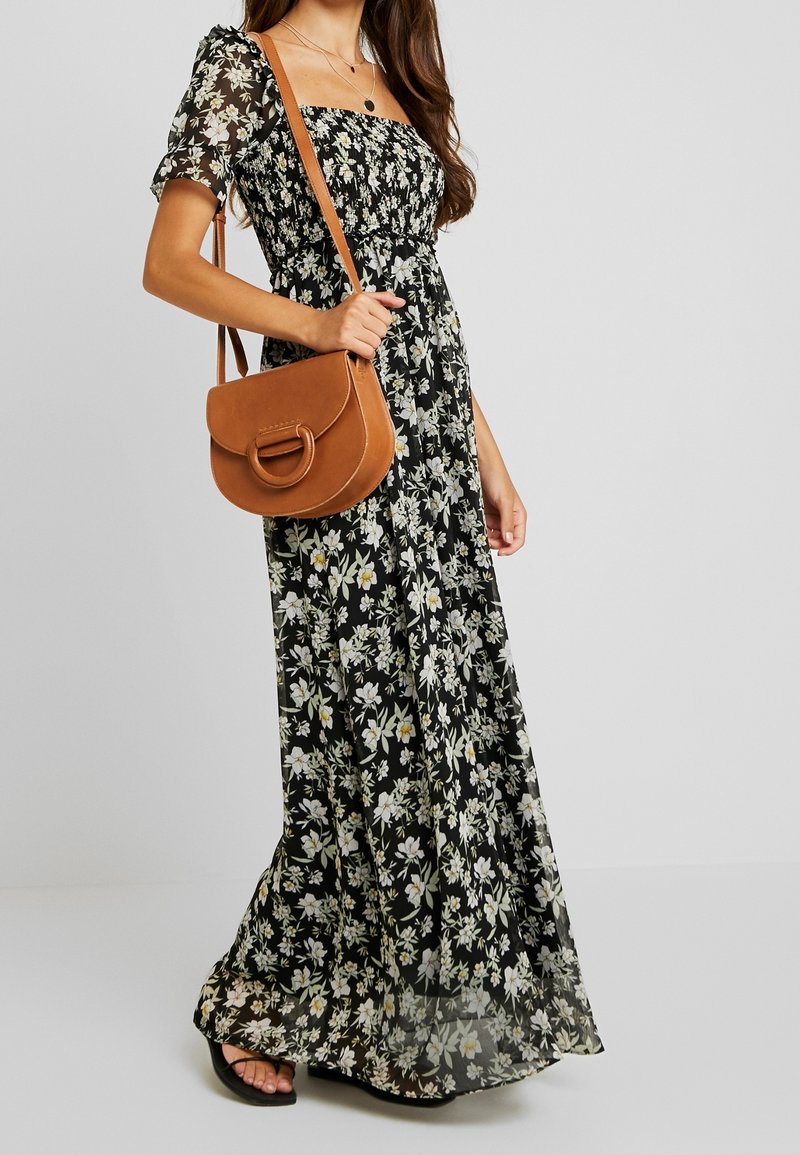 Maxiklänning i svart tyg med vit och gul blommig tryck, korta puffärmar, elastisk bodice, tillsammans med en brun axelremsväska.