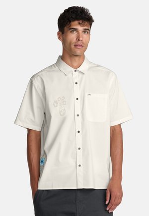 Homme portant une chemise blanche à manches courtes avec poche poitrine et imprimé floral subtil, associée à un pantalon foncé, debout devant un fond blanc.