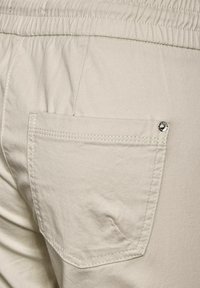 Gros plan sur la poche arrière d'un pantalon beige avec des coutures visibles et un petit rivet en métal dans le coin supérieur droit.