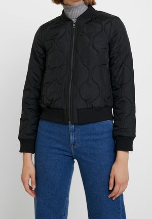 Personne portant une veste matelassée noire zippée sur un col roulé gris et un jean bleu taille haute, debout devant un fond uni.