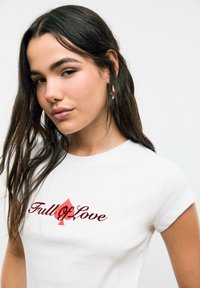 T-shirt en coton blanc avec un texte rouge en cursive "Plein d'Amour" et un graphique de pique rouge. Manches courtes et col rond. Texture lisse.