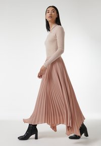 Lichtroze, langslijfs top met een zachte textuur, gecombineerd met een geplooide, asymmetrische roze rok; zwarte, hoge laarzen maken de outfit compleet.