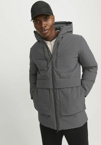 Grå isoleret jakke med hætte, der har et tofarvet design, frontlynlås og sidelommer. Modellen har en sort kasket og en beige top på.