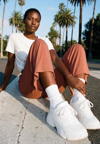 Fiatal személy ül az úton pálmafák között, fehér tornacipőt, fehér zoknit, rózsaszín nadrágot és fehér pólót visel napos időben.