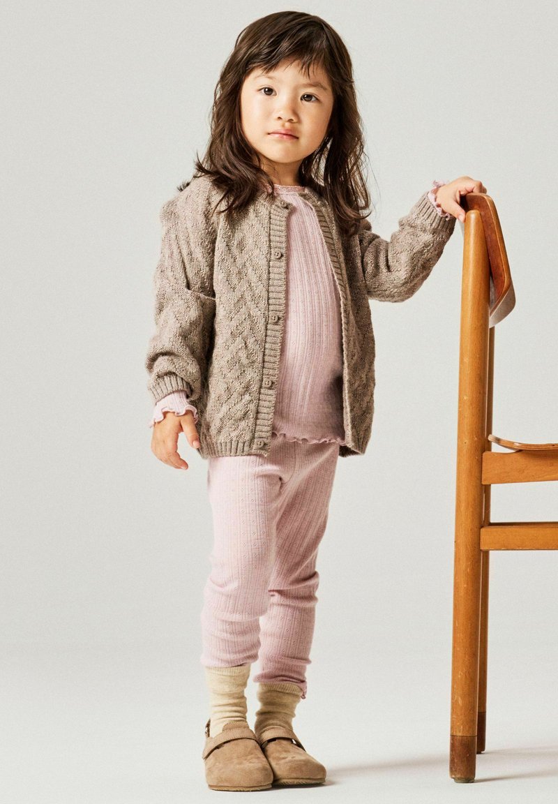 Jeune enfant portant une tenue en tricot rose et un cardigan gris, debout tenant le dossier d'une chaise en bois sur un fond clair uni.