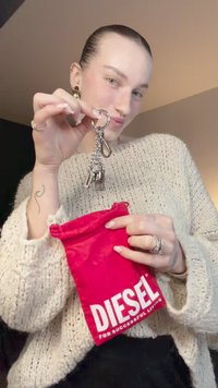 Portachiavi d'argento con più ciondoli, tra cui un lucchetto e una clip, tenuto sopra una pochette rossa con il logo "DIESEL" in lettere bianche. Maglione lavorato a maglia con trama.