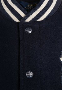 Marinblå ulljacka med randig ribbad krage, metallknappar med "Polo RL"-logotyp och broderade detaljer.