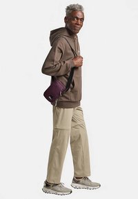 Brown hoodie, beige cargo pants, and gray sneakers. Carrying a small purple crossbody bag. Visible stitching and zippered detail on bag.