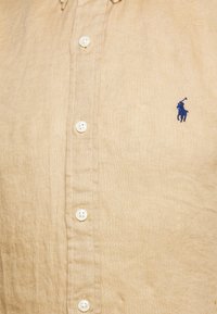 Béžová lněná košile s límcem na knoflíky, s bílými knoflíky a modrým vyšívaným logem na levé straně hrudi. Hladká textura.