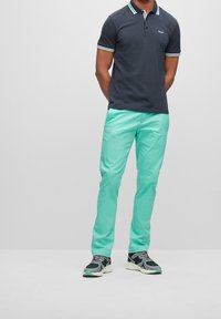 Man i en marinblå polo, mintgröna byxor och svarta, grå och vita sneakers som står framför en enkel ljusgrå bakgrund.