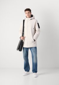 Lys beige hættejakke med lynlås foran, en sort patch på venstre ærme, parret med blå jeans og hvide sneakers. Sort messenger taske.