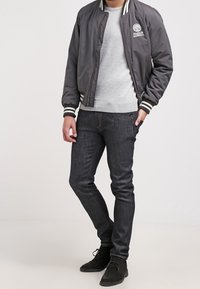 Giubbotto bomber grigio con costine a righe, caratterizzato da un emblema, indossato sopra un maglione grigio chiaro e jeans in denim scuri. Scarpe nere completano il look.
