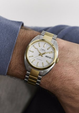 Reloj de pulsera Timex Marlin con correa metálica en oro y plata que muestra la hora 10:11 y la fecha miércoles 14 en una muñeca con vello y una manga azul.