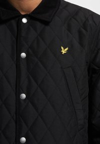 Zwarte gewatteerde jas met een corduroy kraag, zilveren drukknoopjes en een kleine gele adelaar borduursel op de borst. Voorzien van zijzakken.
