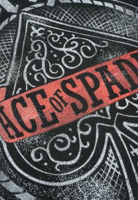 Melns audums ar baltām un sarkanām grafikām, kuras rotā sarežģīti dizaini un teksts "ACE of SPADES" drukātā, nolietotā burtu stilā.
