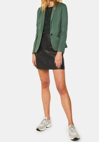 Green blazer over a dark patterned shirt, paired with a black leather mini skirt and light gray athletic sneakers with laces.