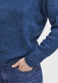 Modrá pletená mikina s jemnou textúrou, s ribovanými manžetami. Kombinovaná s tmavými džínsy, zobrazená s rukou v kapsičke.