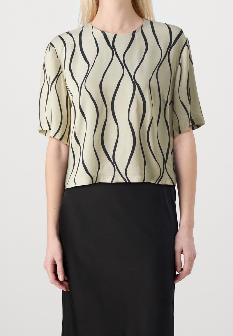 Femme portant un haut beige à manches courtes avec des rayures verticales ondulées noires et une jupe noire taille haute sur un fond neutre.