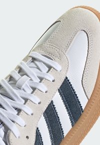 Sneaker, amelynek fehér bőrből készült teste van, szürke velúr részletekkel, sötétkék csíkos anyaggal és gumi talppal. A fűzők fehérek.