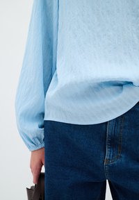 Ljusblå texturerad blus med puffärmar, tillsammans med mörkblå denimjeans. Blusen har en kurvad fåll och subtila vertikala ränder.