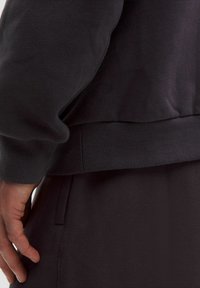 Sort sweatshirt med en struktureret overflade, tætsiddende manchetter og ribbet kant. Logo-detalje synlig nær den nederste søm. Stoffet ser blødt ud.