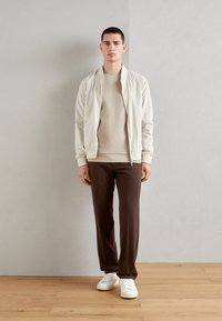 Giacca bomber beige chiaro, felpa beige, pantaloni marroni e sneakers bianche. Design semplice con una vestibilità rilassata e texture liscia.