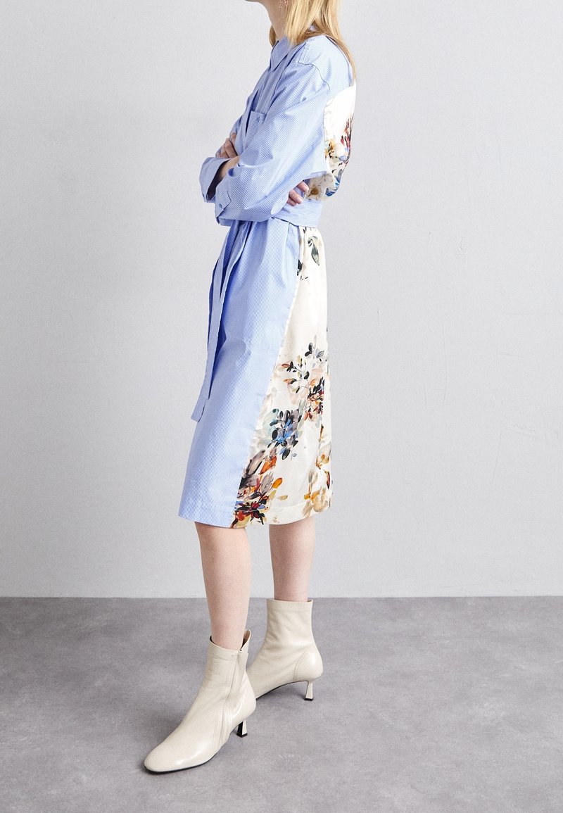 Femme debout, les bras croisés, portant une robe midi bleue à fleurs et des bottines crème sur un fond gris clair uni.