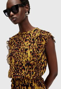 Blouse jaune avec un motif léopard, dotée de manches volantes et d'une texture douce. Le mannequin porte des lunettes de soleil noires surdimensionnées.