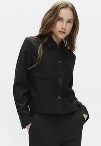 Sort cropped jakke lavet af glat stof, med kraveudskæring, frontknaplukning og to brystlommer.