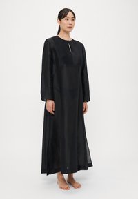 Vestido largo negro de tela ligera, con cuello alto, detalle plisado en el frente y aberturas laterales, con mangas largas.