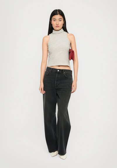 Frau in ärmellosem weißen gestreiften Rollkragenpulli, lockeren schwarzen Jeans, weißen spitzen Schuhen, hält eine rote Clutch unter dem Arm und steht vor einem weißen Hintergrund.