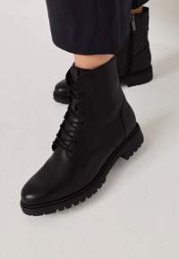 Bottes de cheville en cuir noir avec un bout rond, un laçage à l'avant et une semelle robuste ; elles disposent d'une fermeture éclair sur le côté pour un enfilage facile.