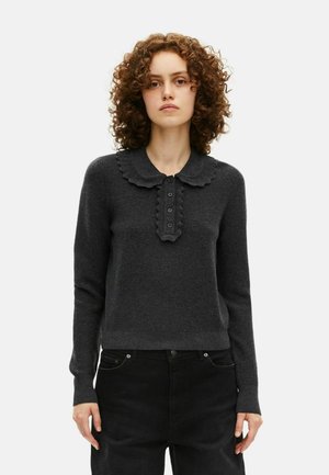 Vrouw met krullend haar, gekleed in een donkergrijze lange mouw trui met een geribbelde kraag en knoopdetails, gecombineerd met zwarte broek.