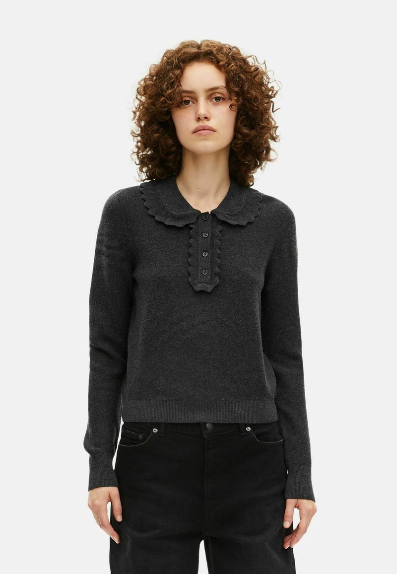 Vrouw met krullend haar, gekleed in een donkergrijze lange mouw trui met een geribbelde kraag en knoopdetails, gecombineerd met zwarte broek.