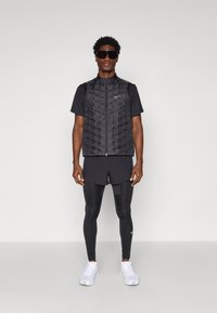 Vesta puffer neagră, cu suprafață texturată, cu fermoar și buzunare frontale, purtată peste un tricou negru și pantaloni scurți; asortată cu jambiere negre și adidași albi.