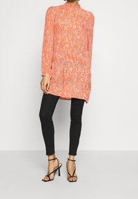 Femme portant un tunique à manches longues florale corail avec un col volanté, un pantalon skinny noir et des sandales à talons hauts noires à lanières, debout.