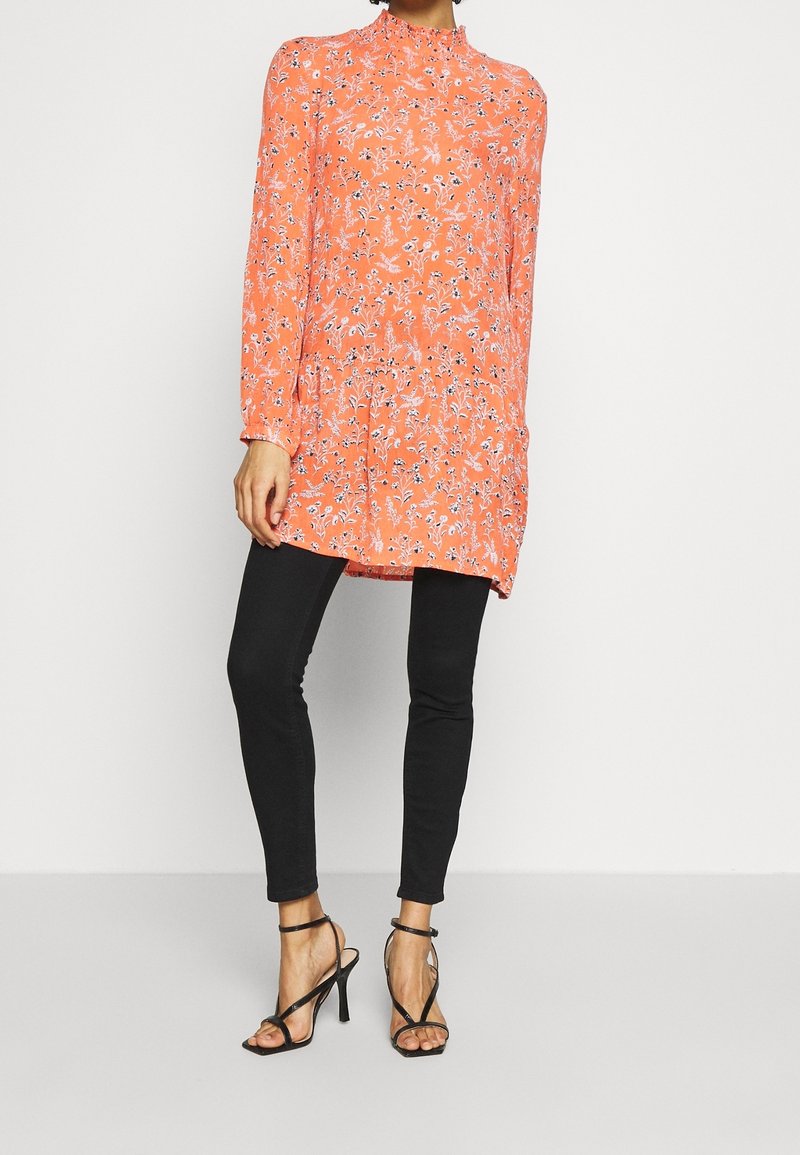 Femme portant un tunique à manches longues florale corail avec un col volanté, un pantalon skinny noir et des sandales à talons hauts noires à lanières, debout.