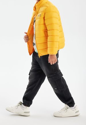 Veste matelassée jaune, pantalon cargo noir avec poches, et baskets blanches avec des accents gris.