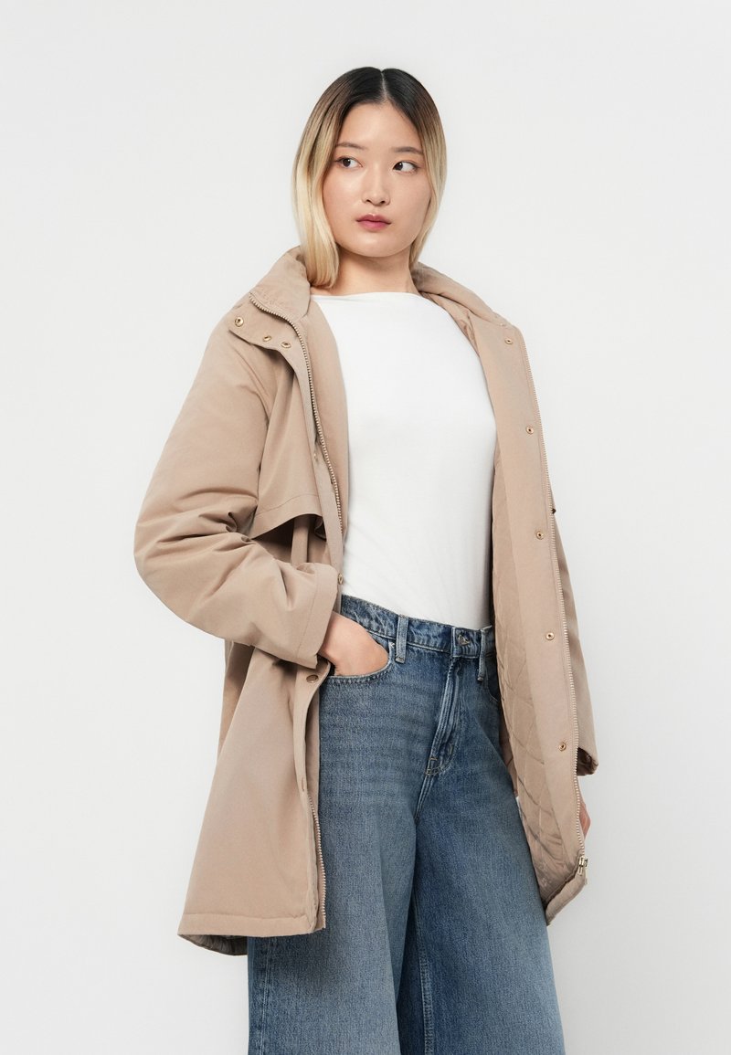 Beżowa kurtka z kapturem, z zamkiem błyskawicznym i napami, pikowanym podbiciem, bocznymi kieszeniami oraz luźnym krojem. Noszona na białej bluzce i dżinsach.