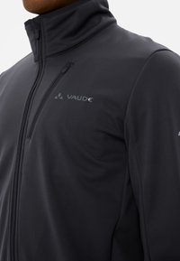 Giacca softshell nera con colletto alto, tasche con zip e accenti del logo ricamati. Tessuto liscio con design aderente.