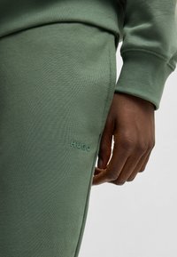 Groene joggingsbroek met geribde boorden, voorzien van een gladde textuur en de omhoog gedrukte merknaam "HUGO" op de linker bovenbeen.