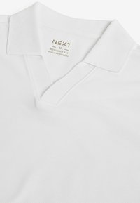 Biały polo z teksturowanej tkaniny, z klasycznym kołnierzykiem i wycięciem w kształcie litery V. Metka informuje o "REGULAR FIT NEXT" wyprodukowane w Bangladeszu.